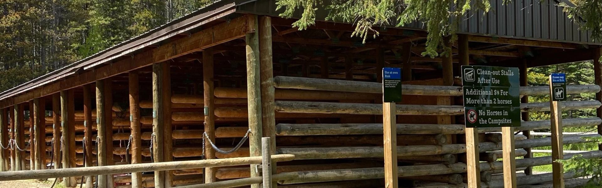 Wooden horse stable with log walls, fenced walkway and green instructional signs outside.