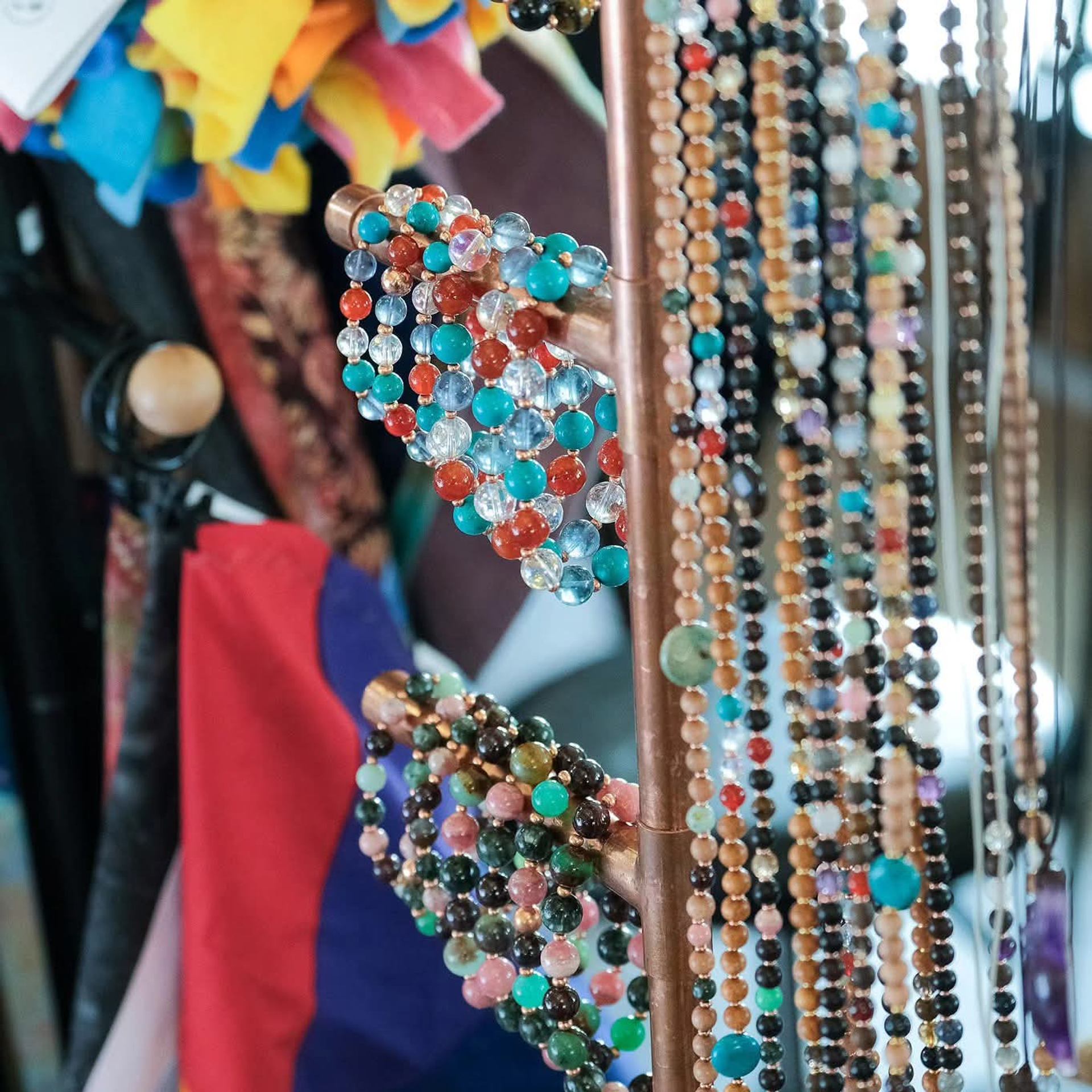Colorful beaded necklaces hang on a rack with patterned fabrics in the background.