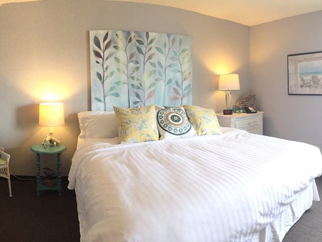 Cozy bedroom with a white bed, colorful pillows, and a leafy painting above, lit by soft lamps.