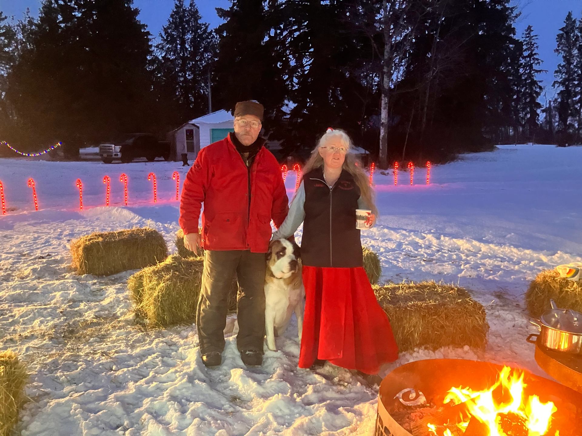 Two people and a dog stand by a snowy campfire with holiday lights behind.