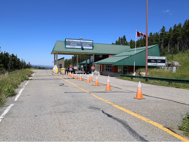 Border checkpoint with green roof, cones, and Canadian flag near trees.