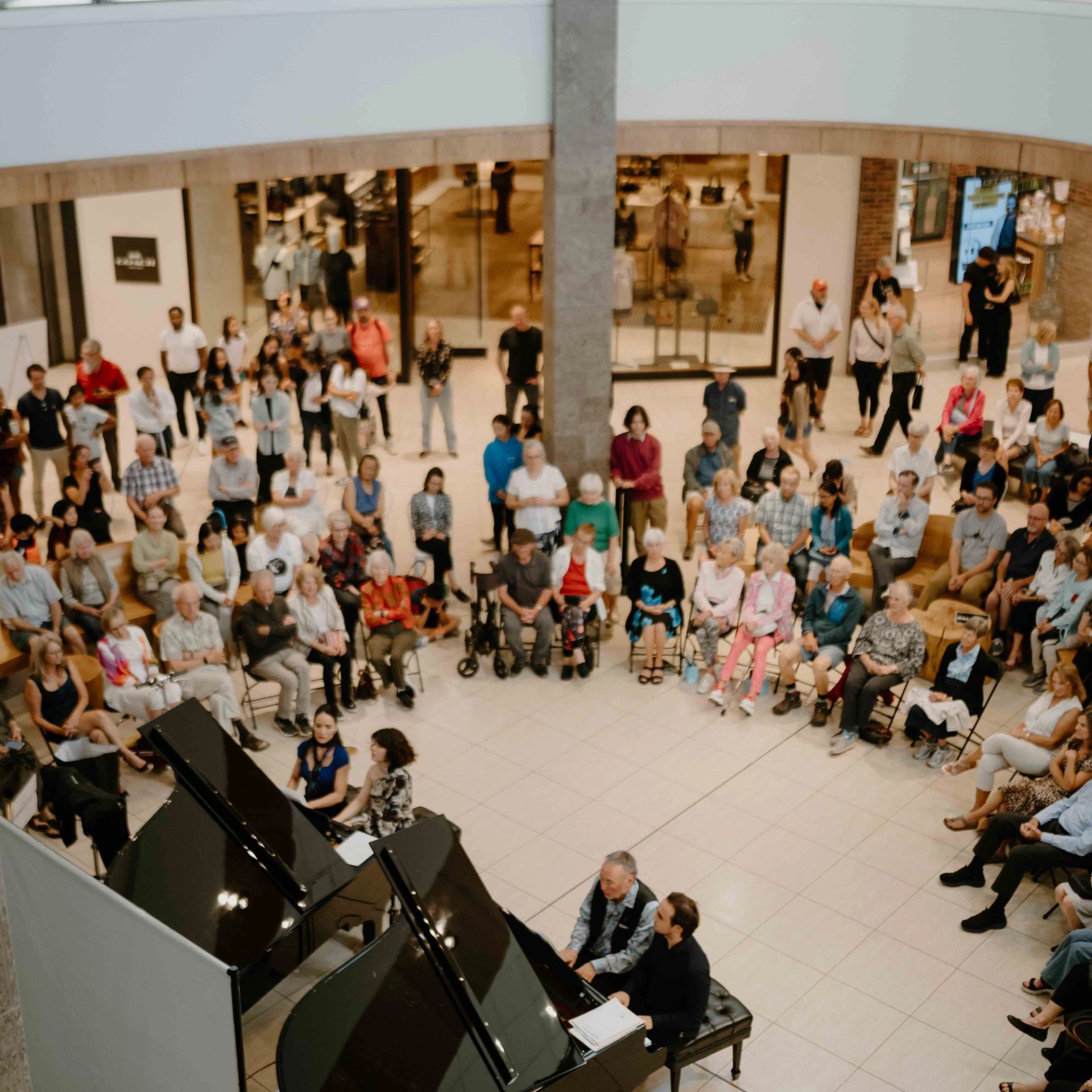 Crowd gathers around two grand pianos in a public indoor space.