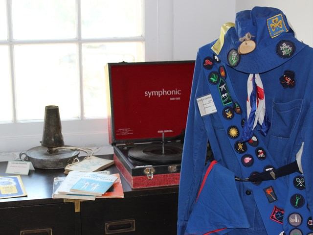 A vintage blue scouting uniform with colorful badges is displayed next to an open red record player and books, suggesting a nostalgic theme.