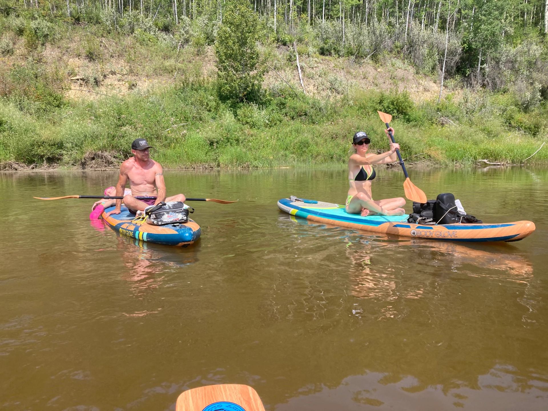 Two people sit on paddleboards in calm river water near a green, wooded shoreline.