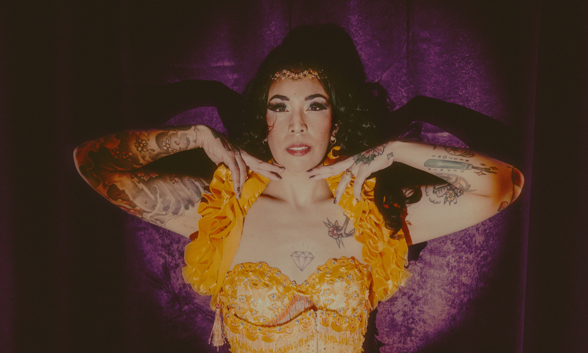 Tattooed performer in yellow ruffled costume poses with hands framing face before purple curtain.