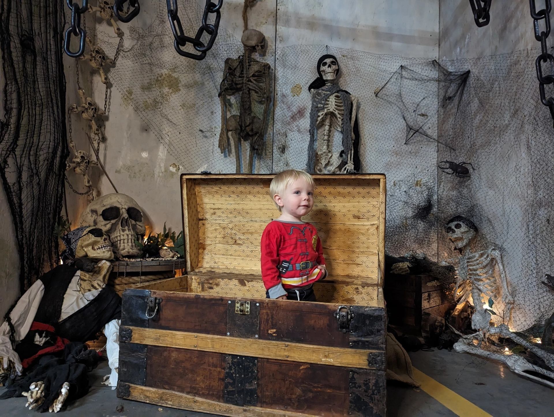 Child in red shirt stands in spooky chest amid Halloween decorations and skeletons.