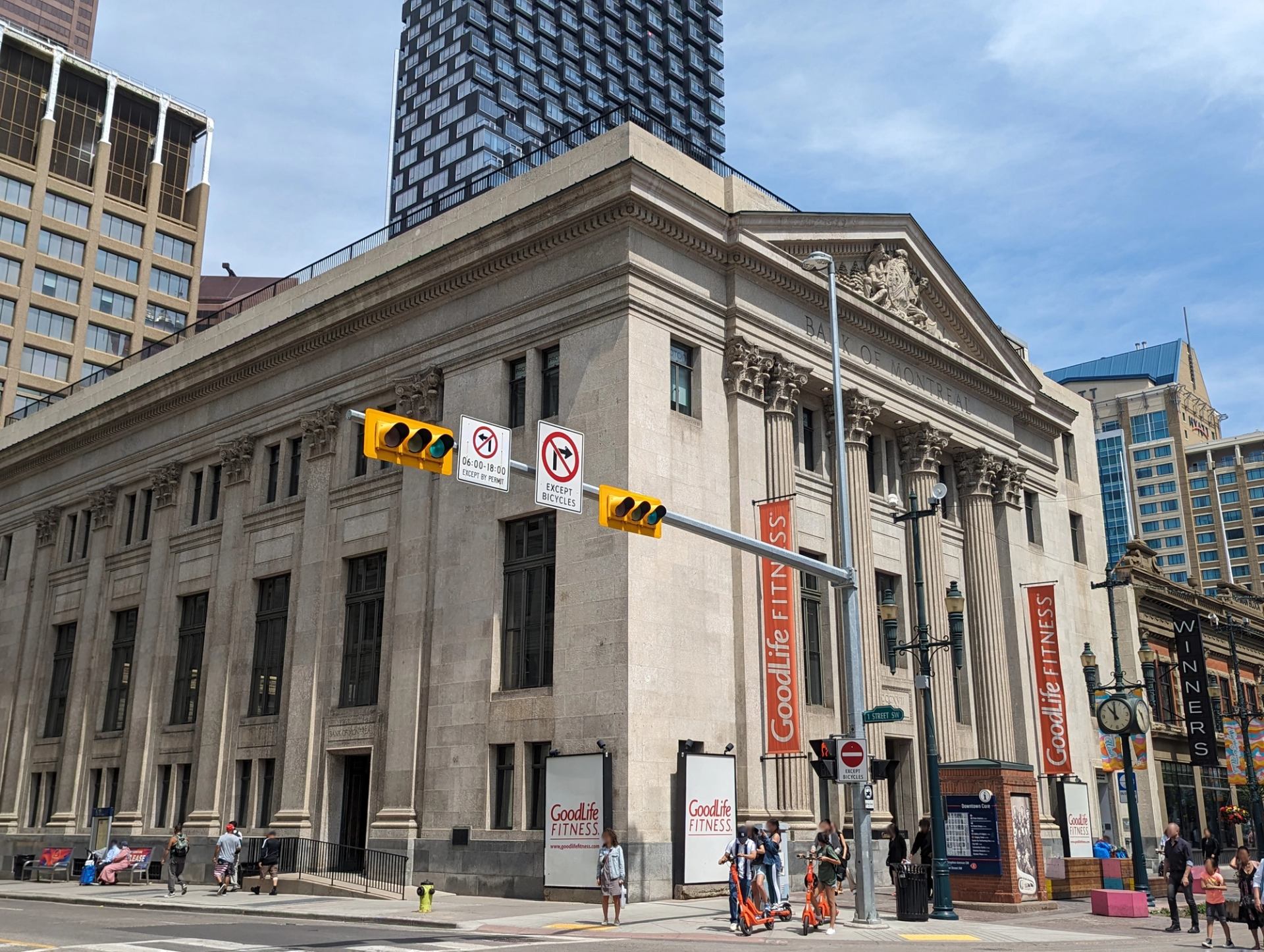 Historic stone bank building with GoodLife Fitness signs at busy downtown intersection.