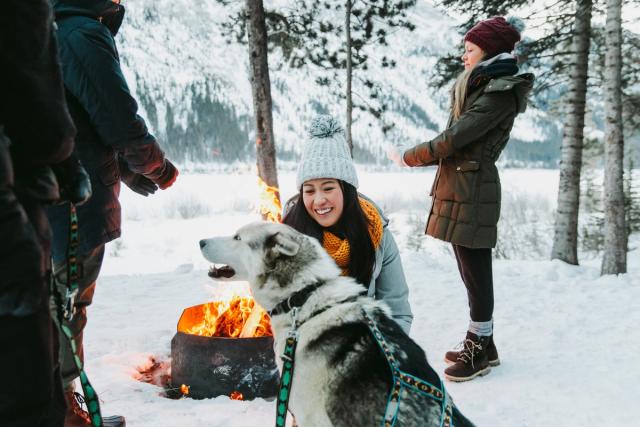 Two people stand around an outdoor fire while one person pays attention to one of the lead huskies, after a dog sled adventure in Spray Lakes in Canmore.