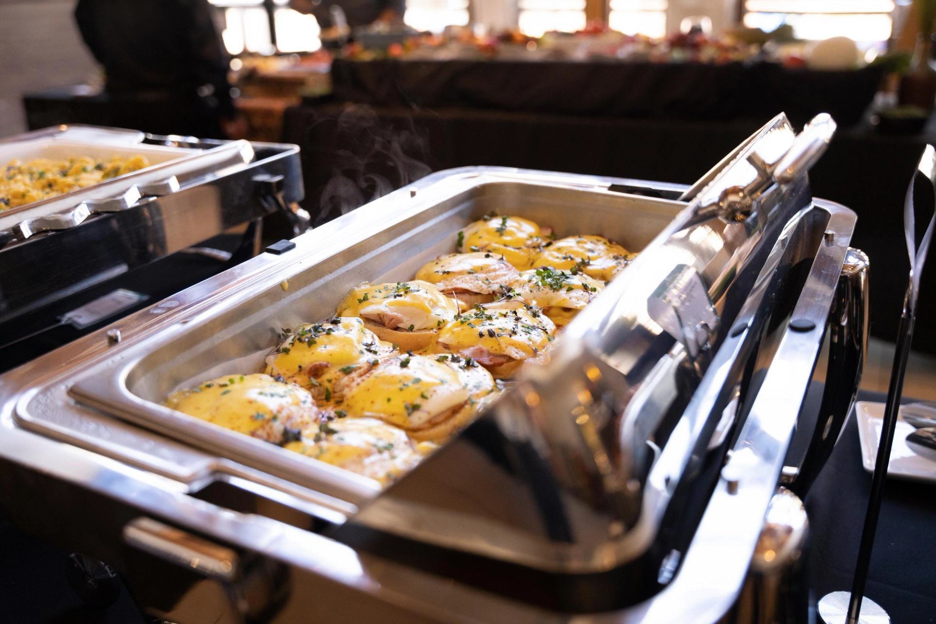A chafing dish filled with steaming Eggs Benedict at a buffet.