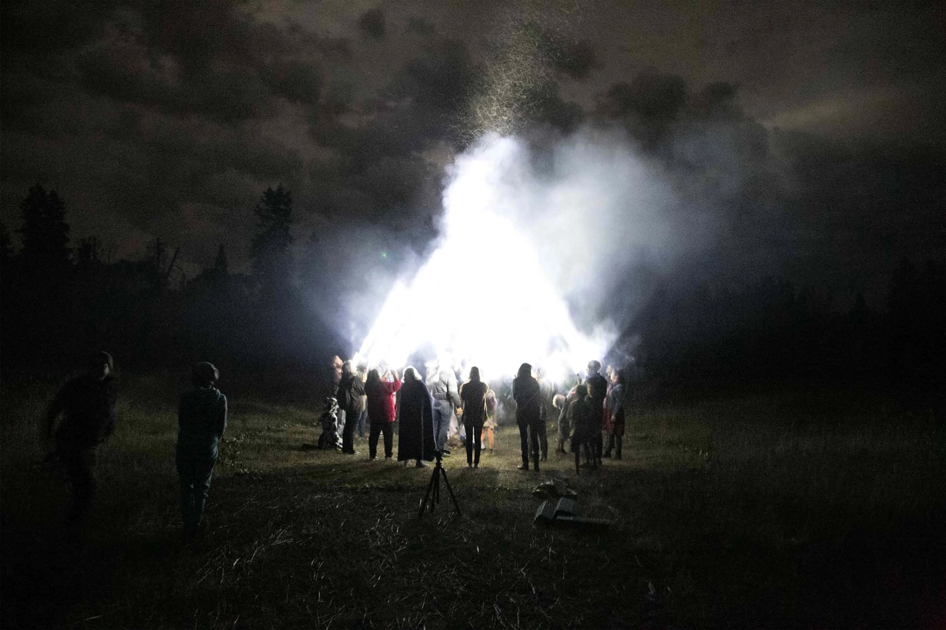 A group of people gathered around a brilliant, smoke-emitting light in a dark field at night.