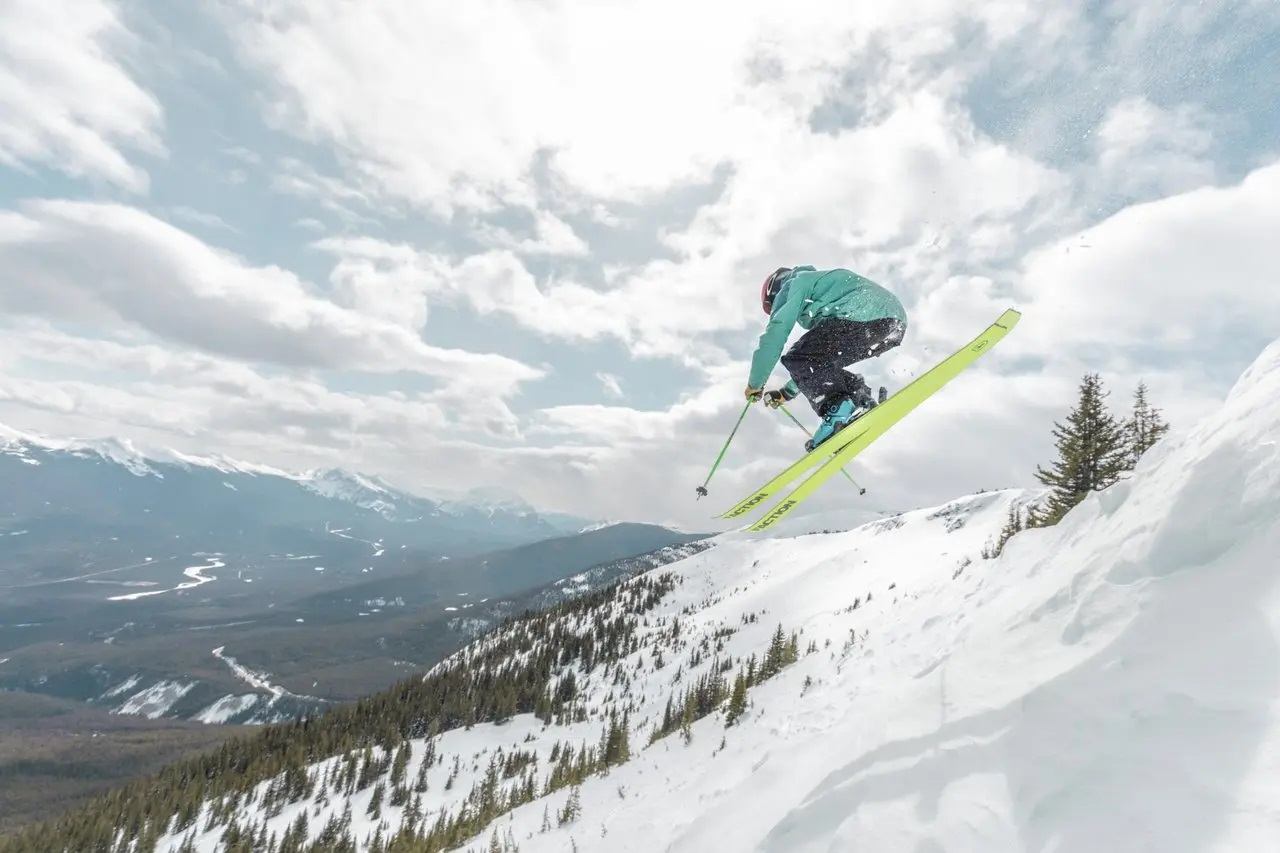 Skier launches off a snowy slope with mountains and cloudy sky in the background.