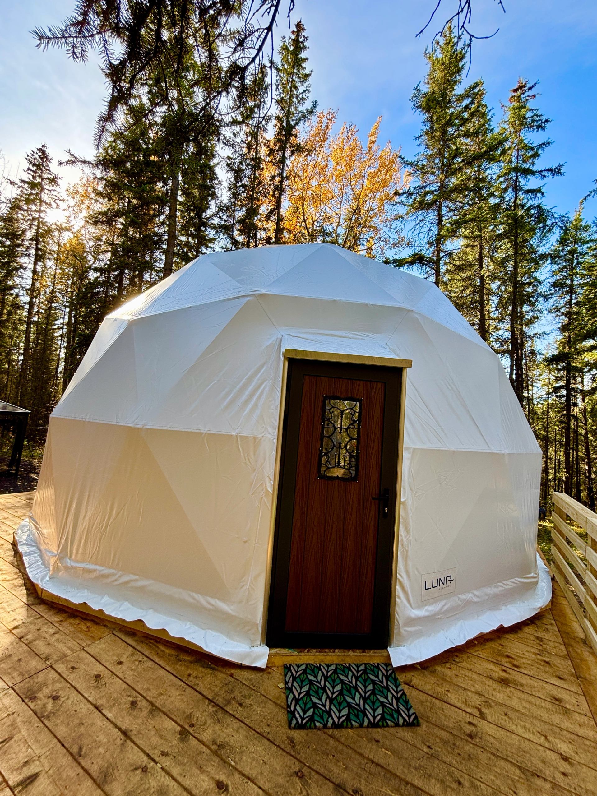 White geodesic dome with wooden door on deck, surrounded by tall evergreen trees.