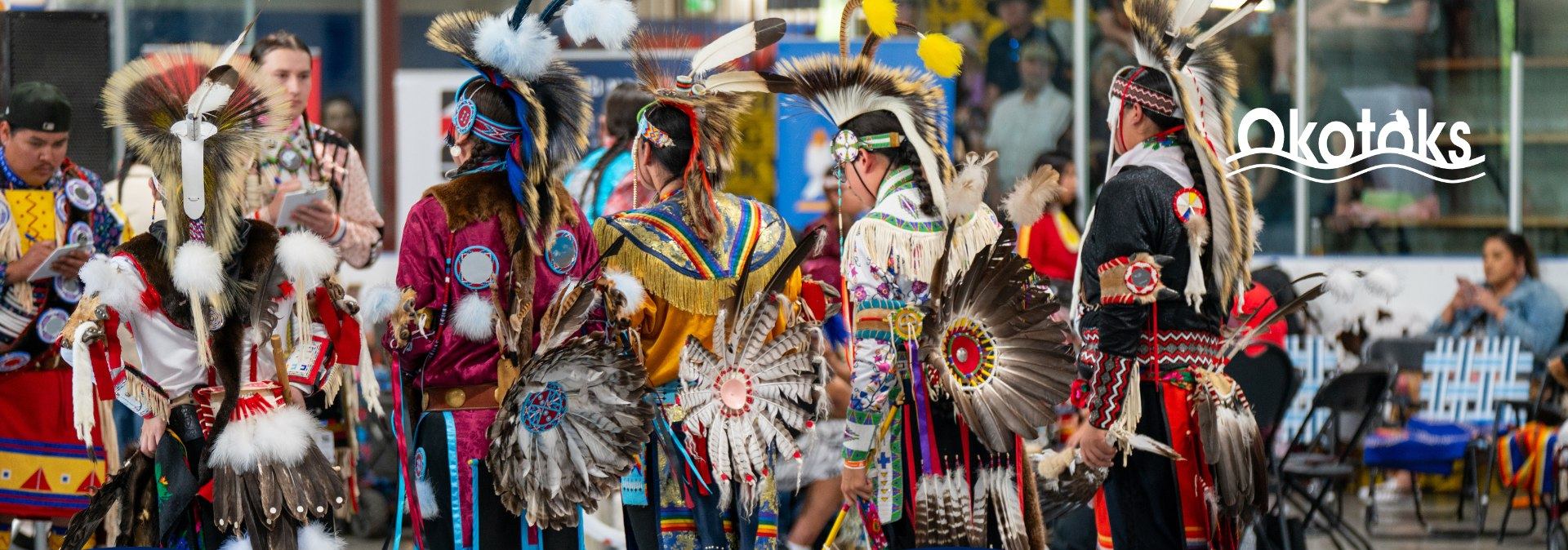 Powwow dancers in colourful regalia with feather bustles, sponsor logos displayed below.