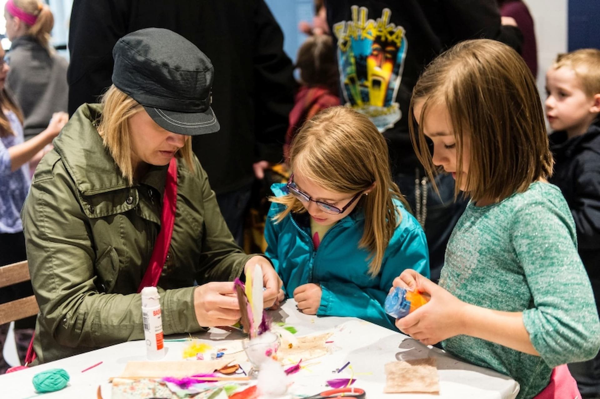 Adults and children sit at a crowded table working on colourful craft projects.