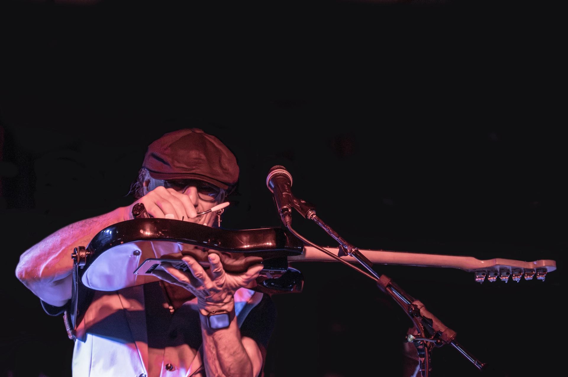 Musician playing an electric guitar on stage, adjusting the instrument near a microphone under dramatic lighting.