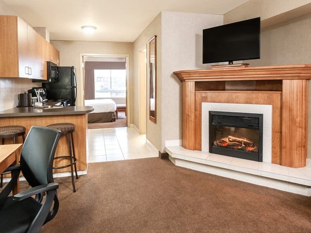 Living area at Canalta Airdrie with fireplace, TV, kitchen, and view into bedroom.