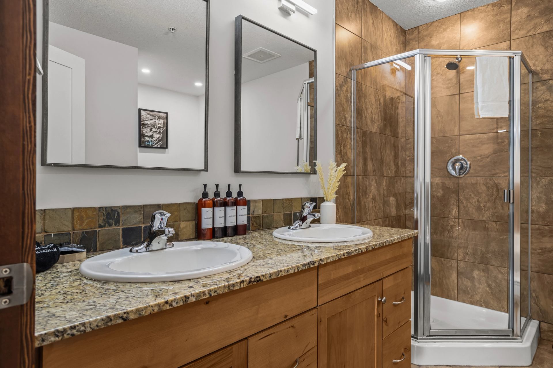 Bathroom with double sinks, granite countertop, glass shower and brown tiled walls.