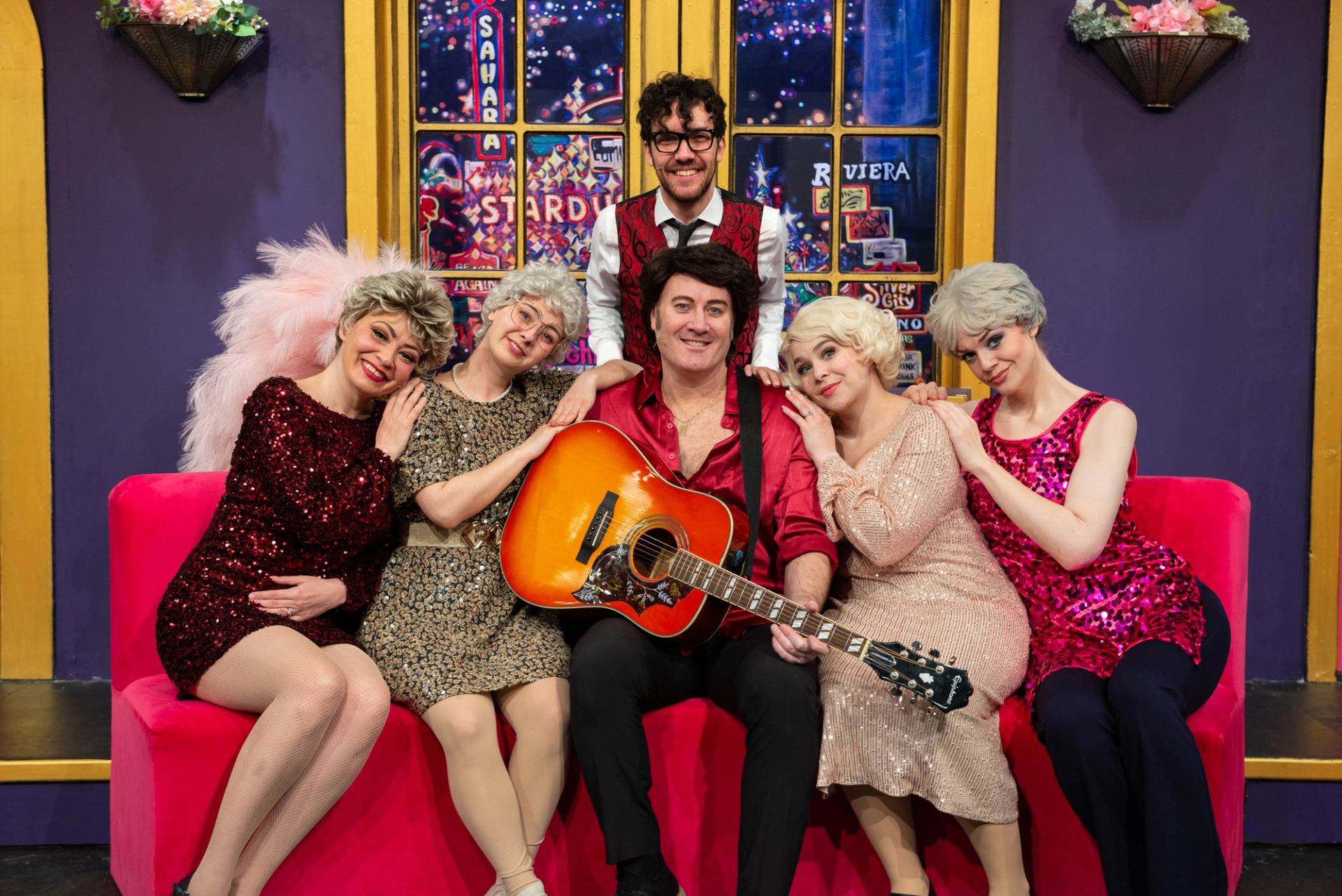 Stage performers in sequined outfits sit on pink couch, man holding guitar, colourful backdrop.