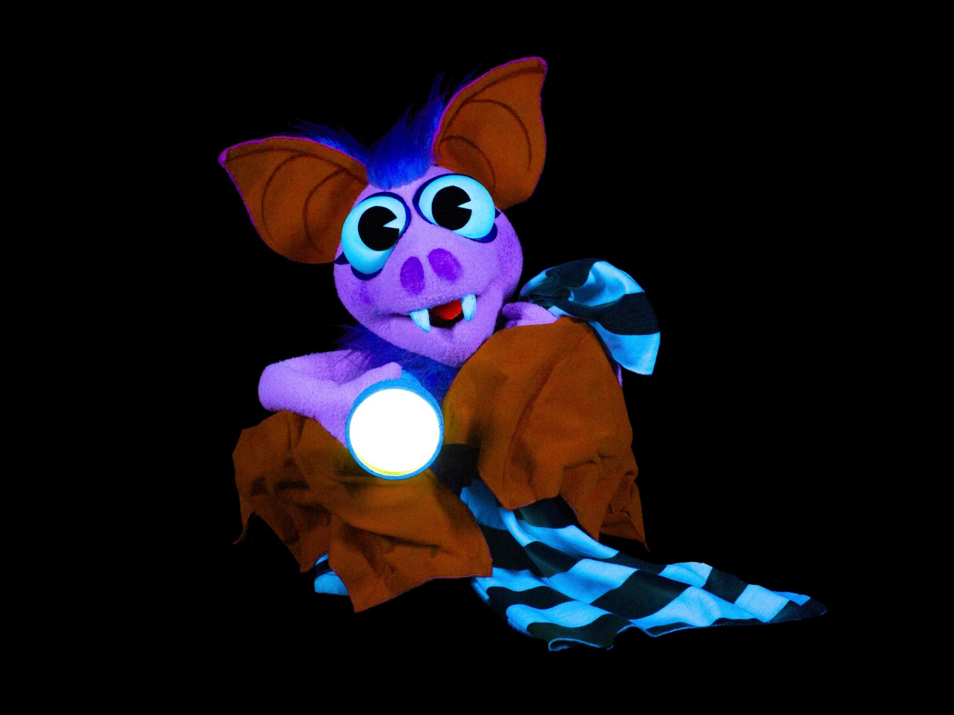 Purple bat puppet holds glowing flashlight and striped blanket against black background.