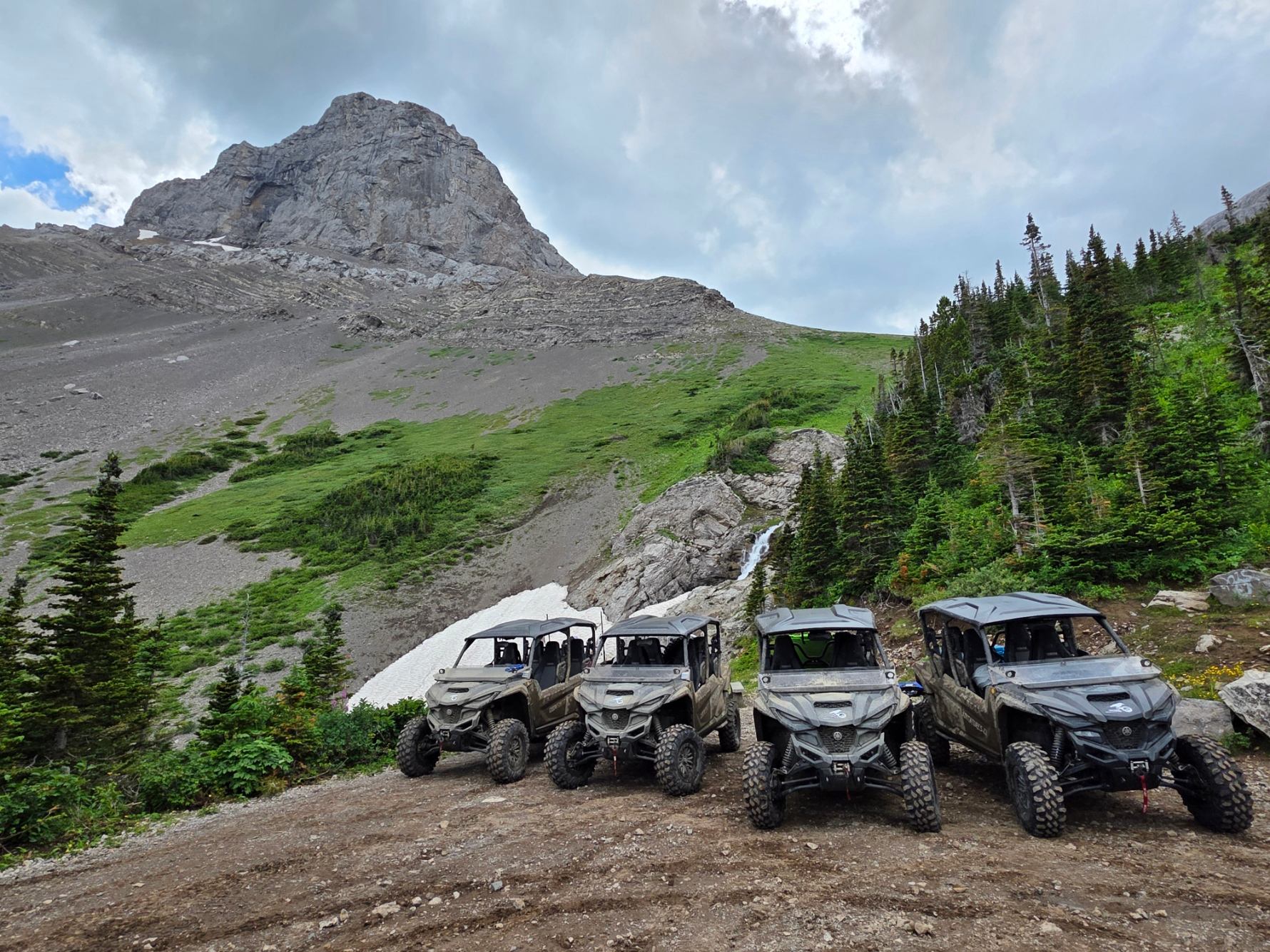 Off-road UTVs parked in front of a rocky mountain and evergreen trees.