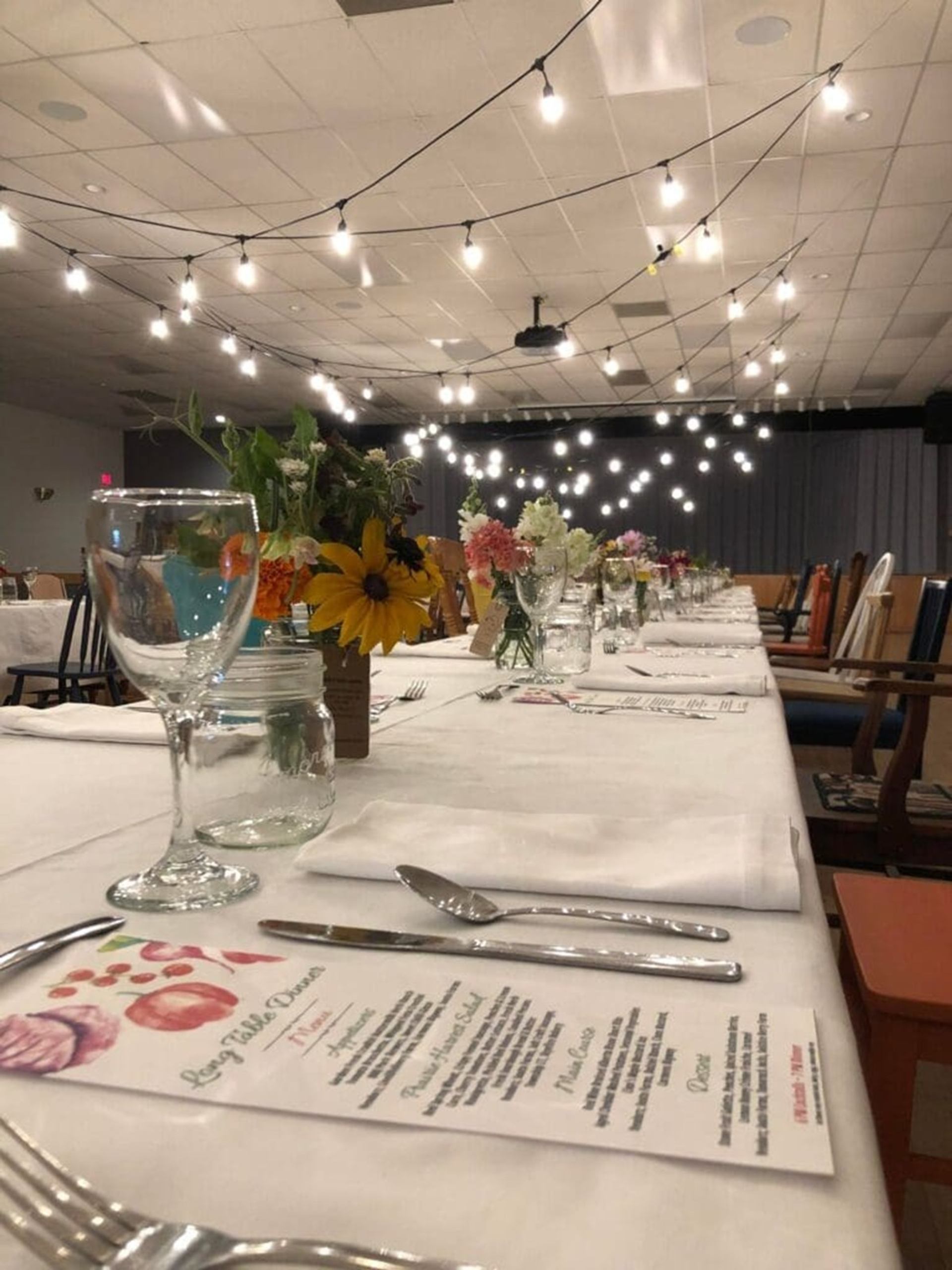 Long banquet table with menus, flowers and glassware under hanging string lights indoors.