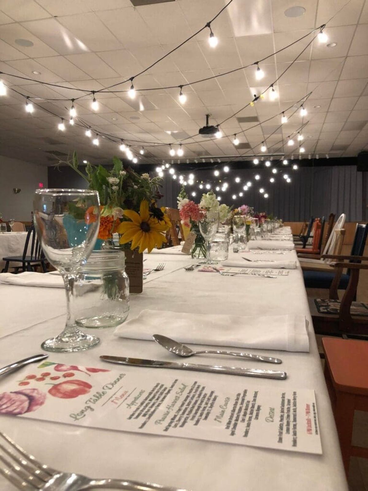 Long banquet table with menus, flowers and glassware under hanging string lights indoors.