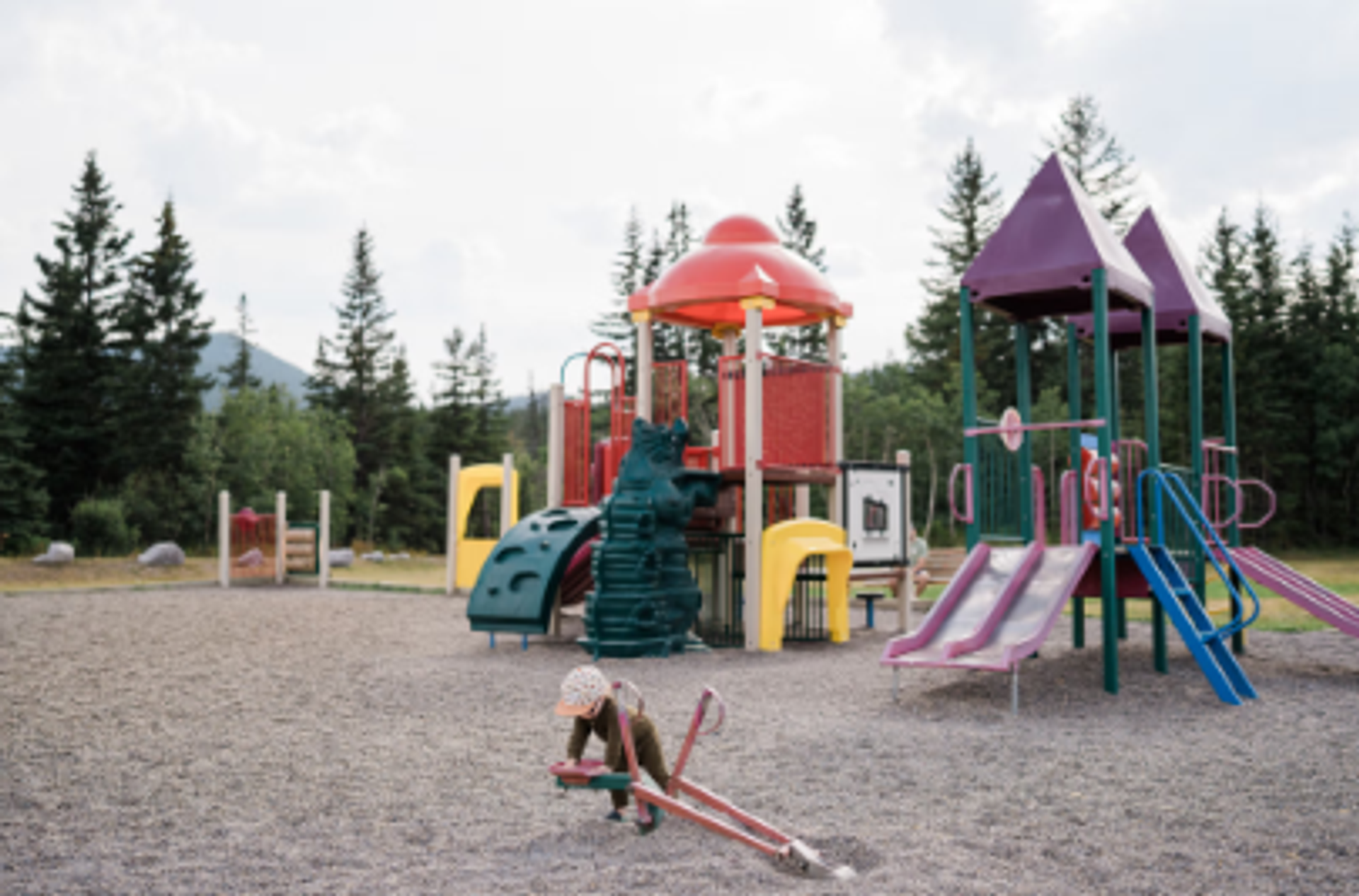 Playground with colourful slides and climbing structures surrounded by trees and distant hills.