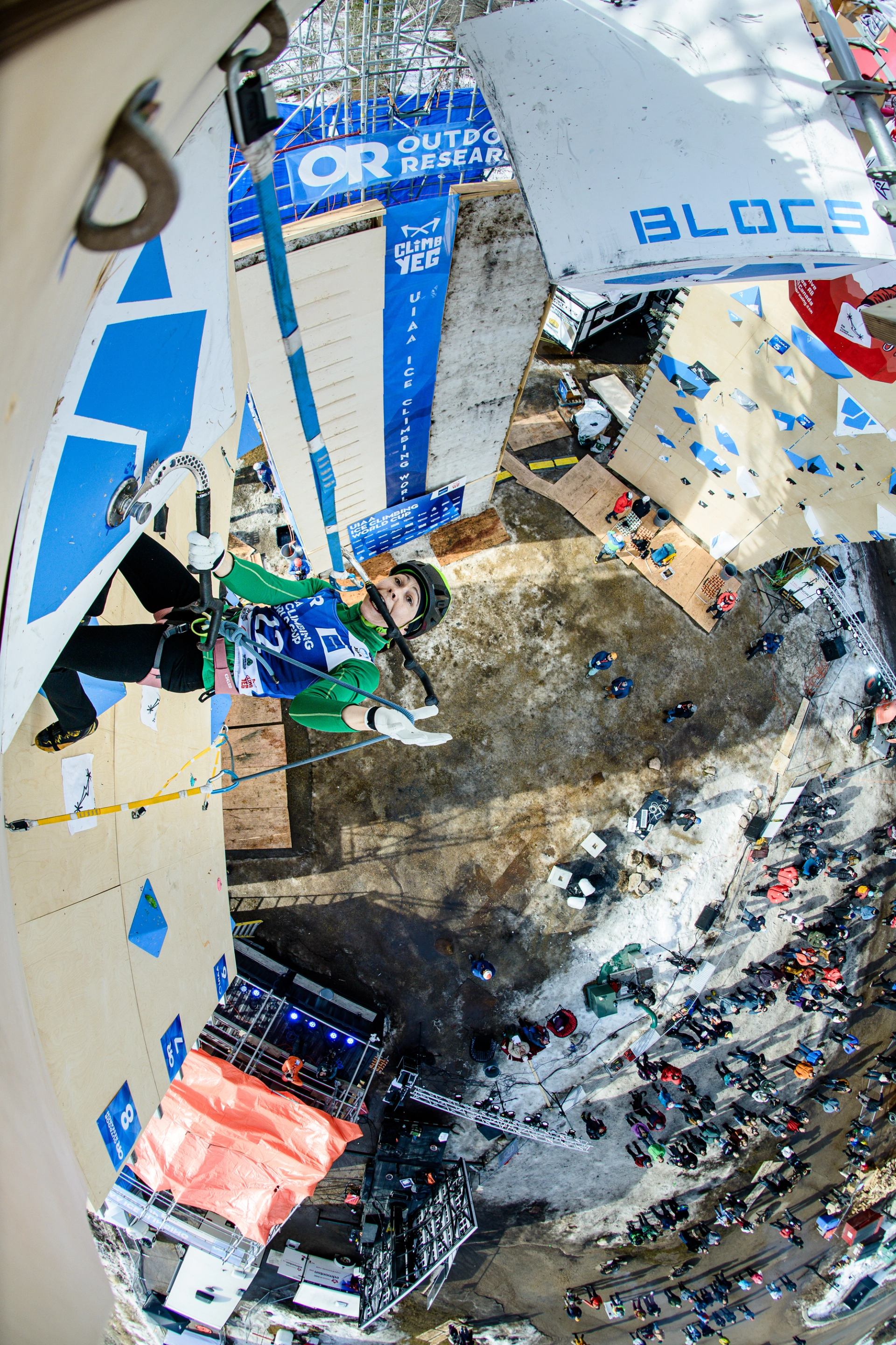 Climber ascending a competition wall with ice tools during an outdoor event.