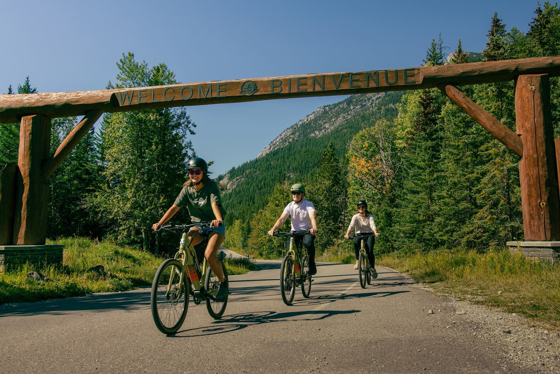 Cyclists riding under a wooden welcome arch surrounded by forest in Banff. 