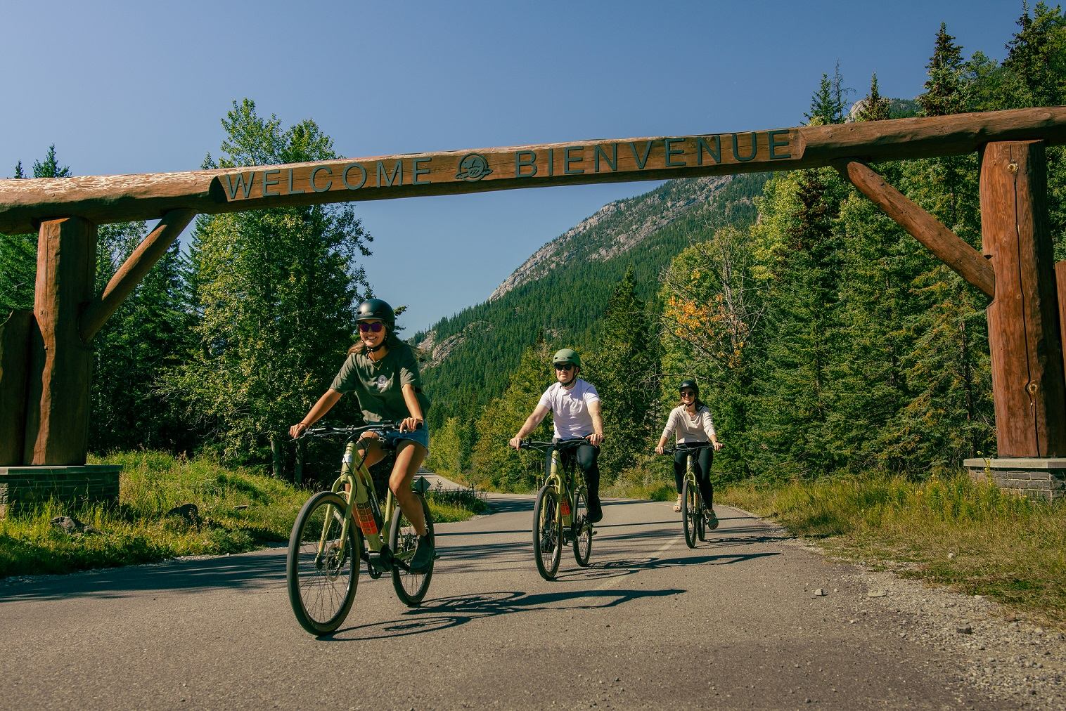 Cyclists riding under a wooden welcome arch surrounded by forest in Banff.