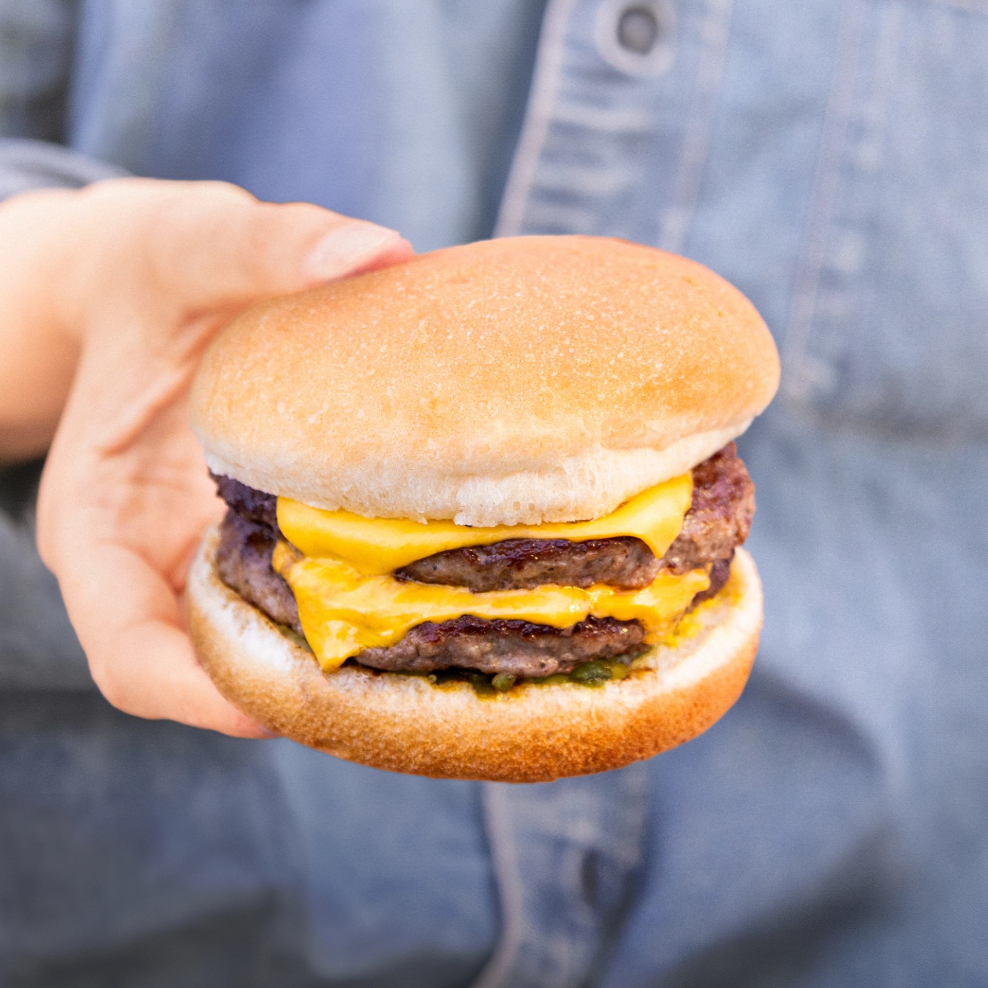 Hand holding a cheeseburger with a bun, yellow cheese, and burger patty in focus.