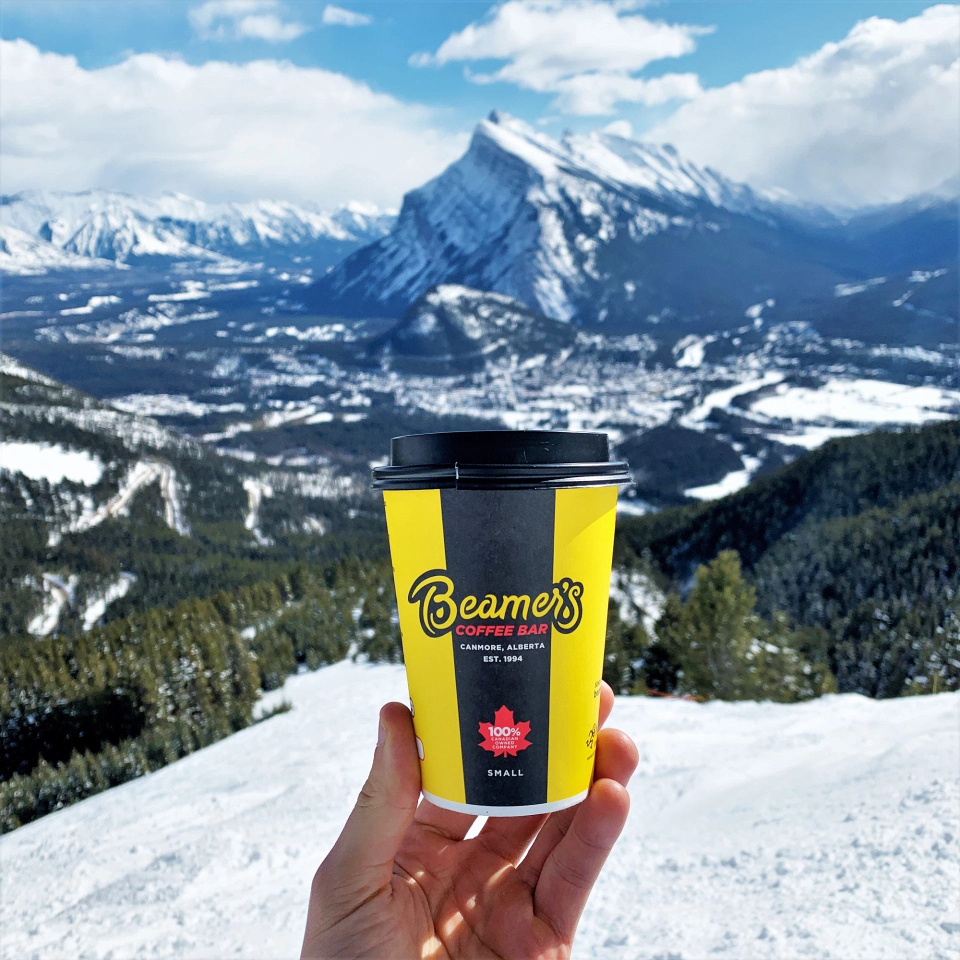 Hand holding a Beamer’s Coffee Bar cup with snowy mountains and a valley landscape in the background.