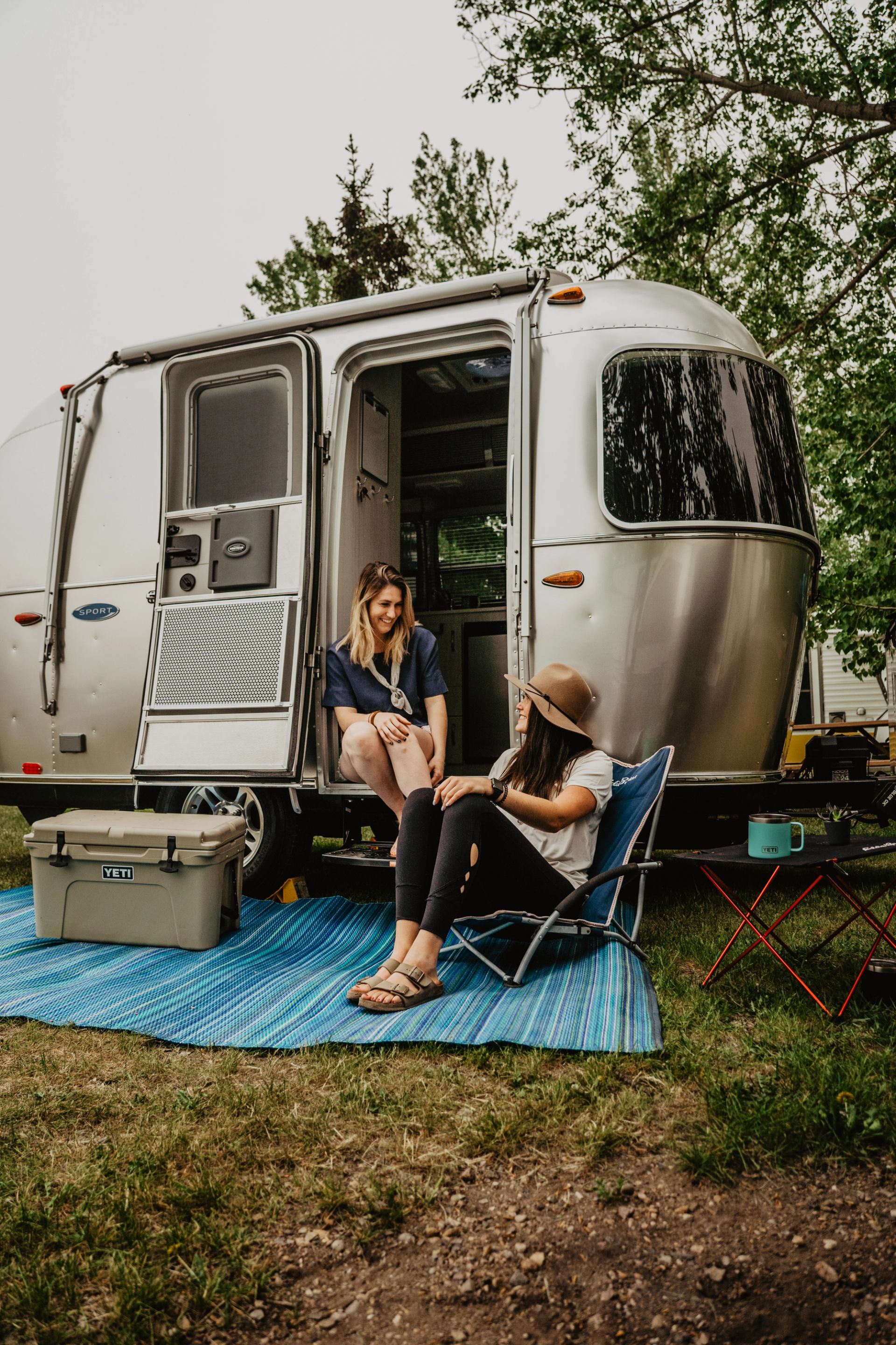 Camping trailer with two people relaxing on outdoor chairs beside cooler and table.
