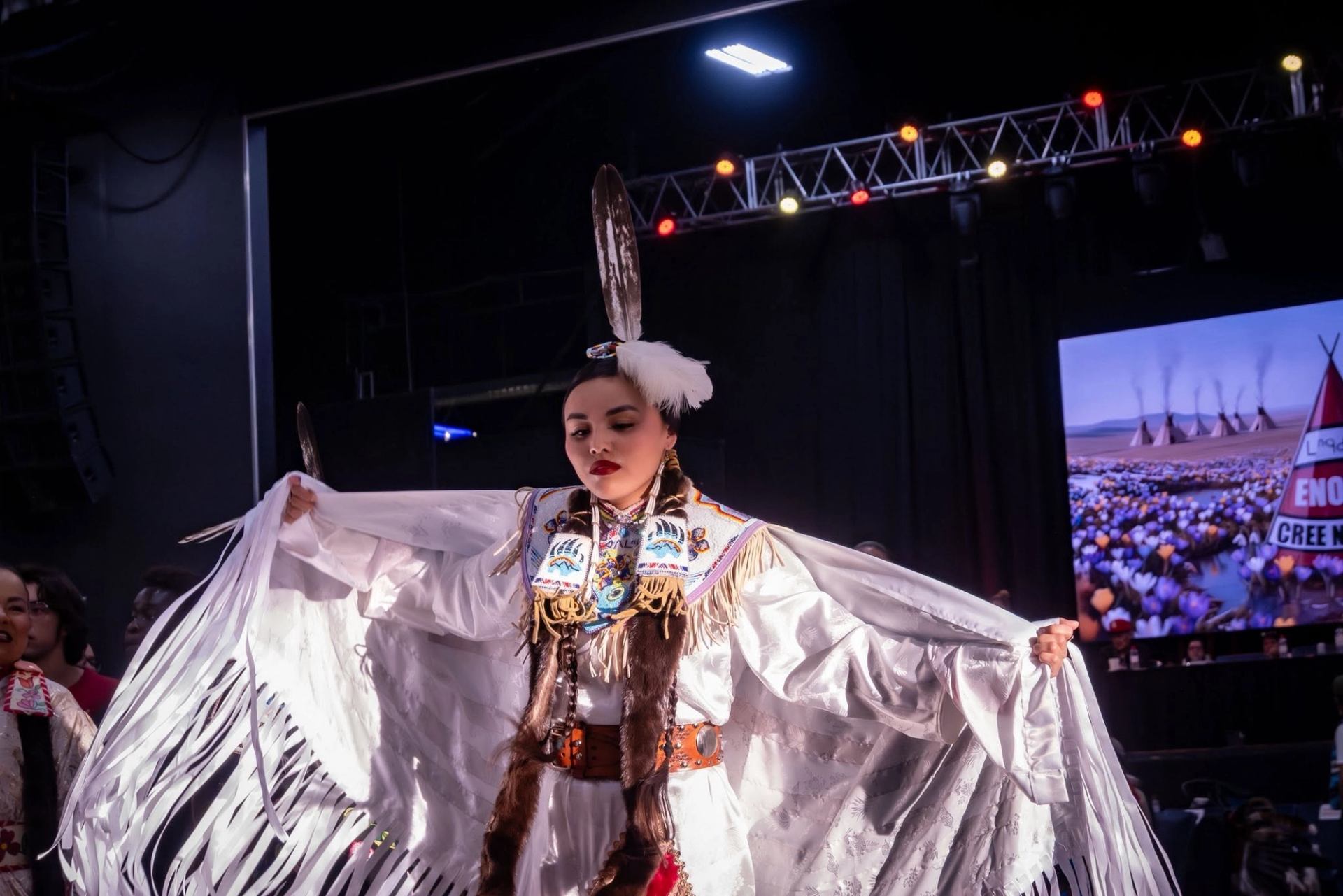 Dancer in white regalia spreads fringed shawl on indoor stage with screen backdrop.