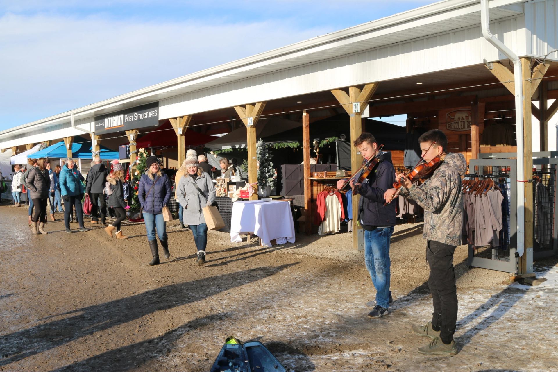 Outdoor market pavilion with musicians playing fiddles beside vendor stalls and shoppers walking pas.