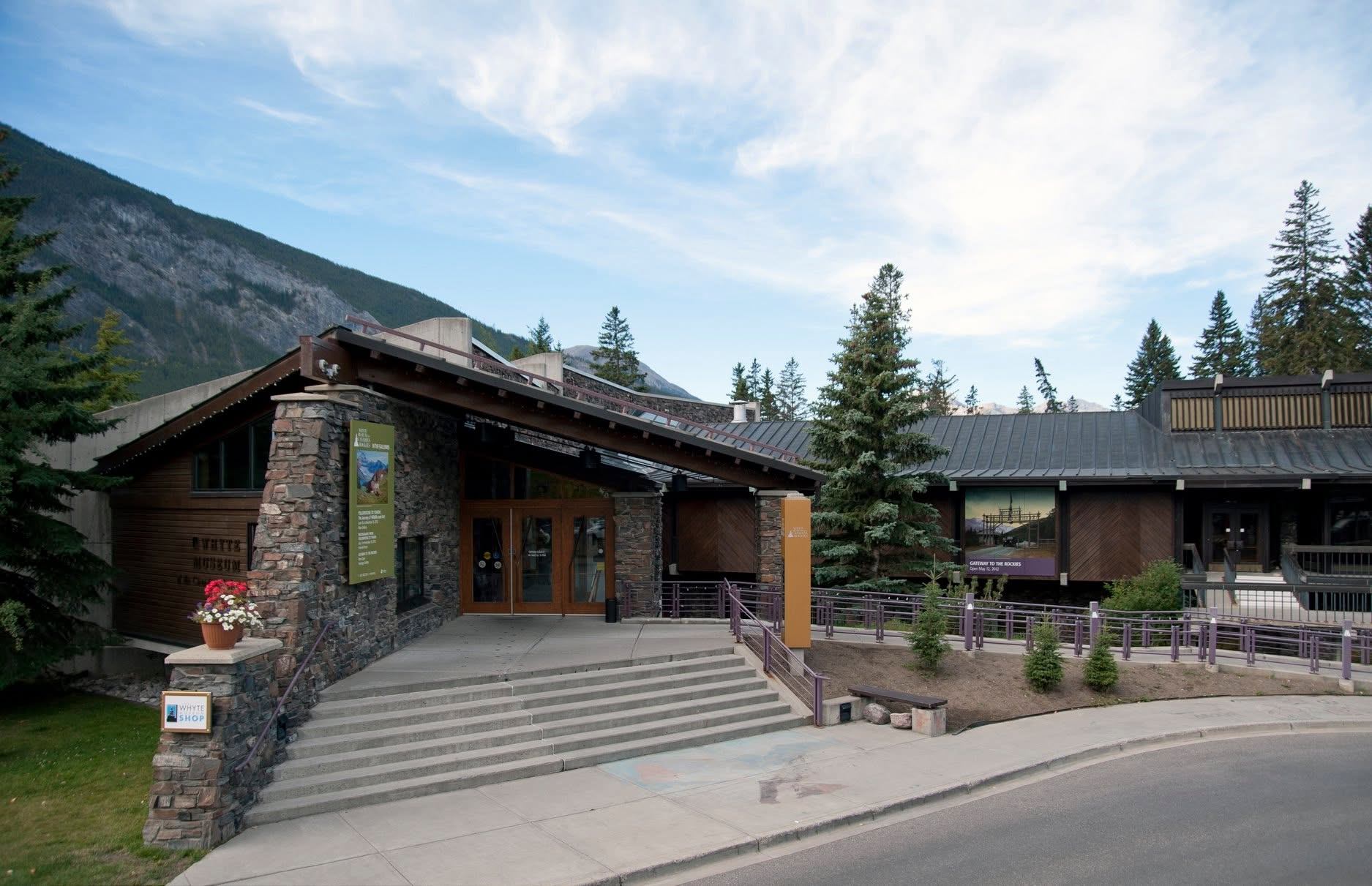 Stone and wood museum entrance with steps, evergreen trees and mountains in background.