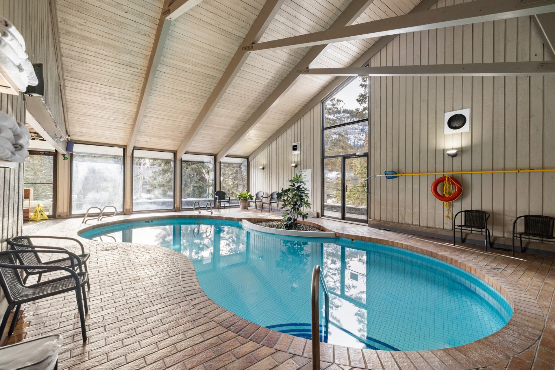 Indoor pool with vaulted ceiling, brick flooring, and large windows with greenery views.
