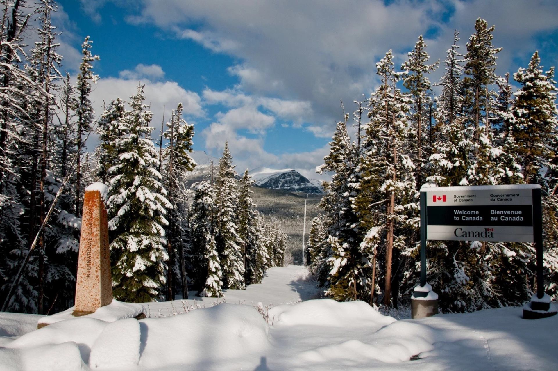 Snowy road between pine trees with International Boundary marker and Welcome to Canada sign.