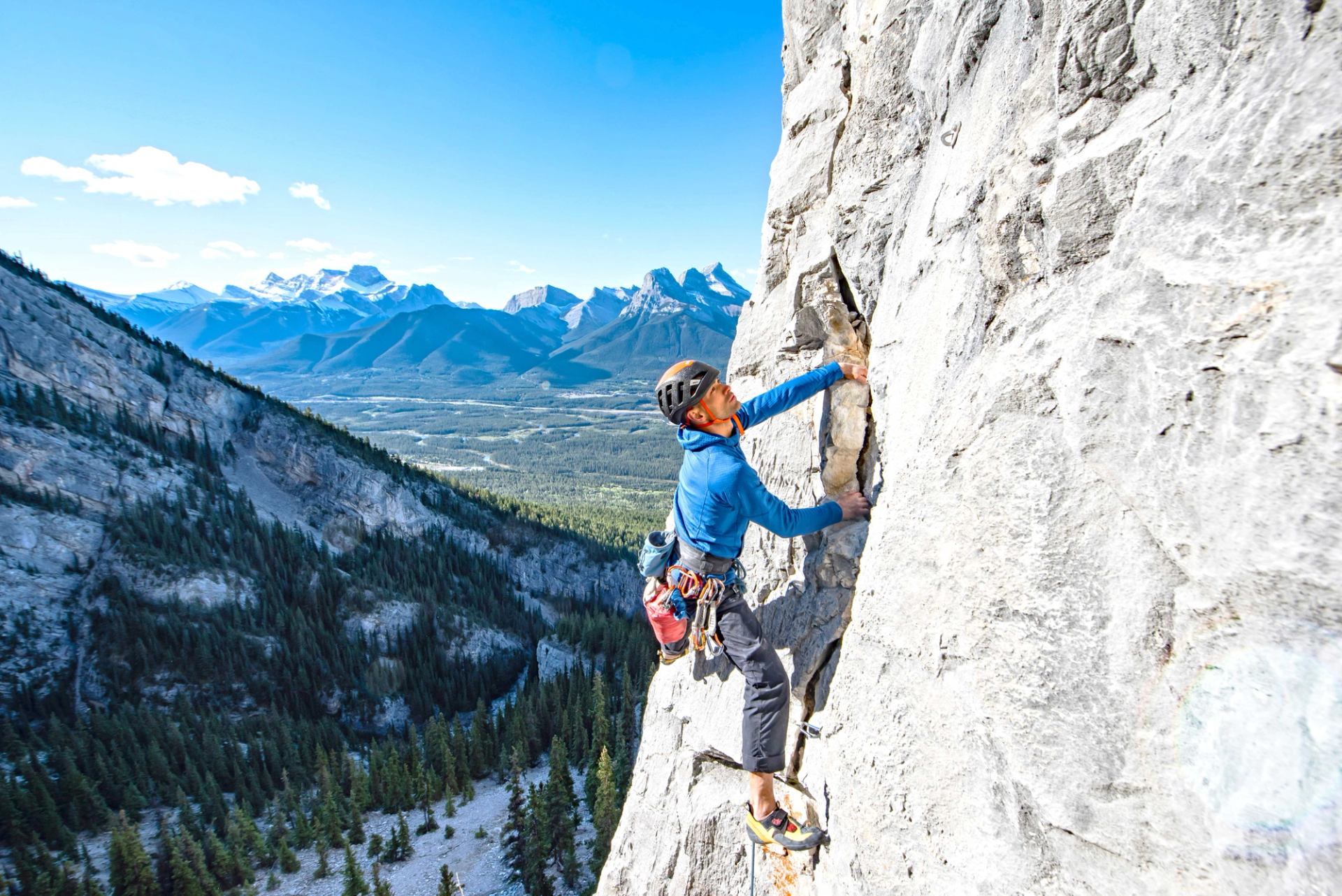 A climber ascends a grey rock face, with a wide valley of pine trees and distant snow-capped mountains visible under a blue sky.