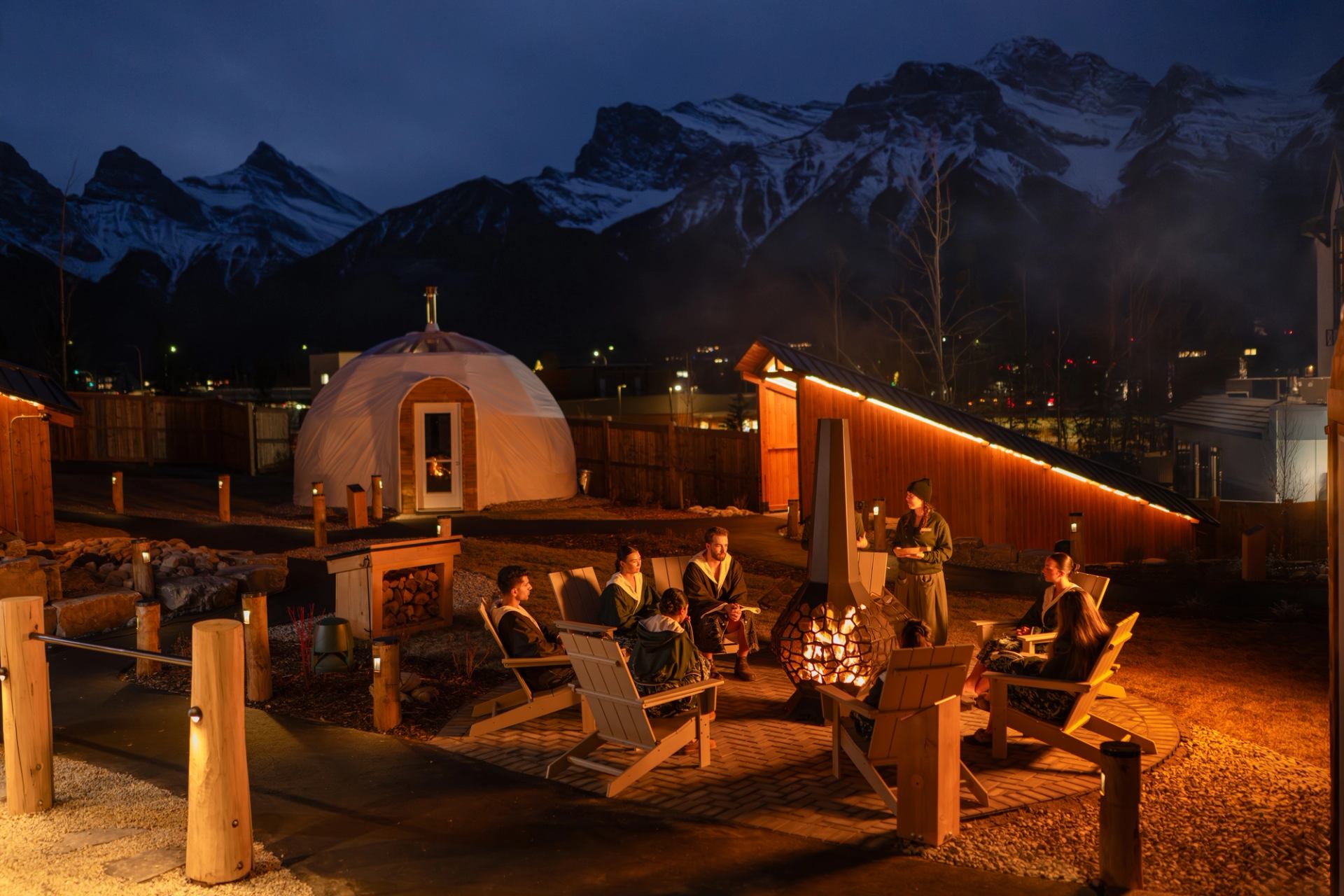 Group gathered around a firepit at Everwild Canmore Nordic Spa with glowing lights and mountain backdrop.