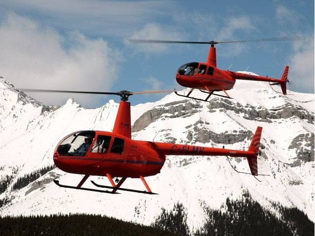 Two red helicopters flying over snow-covered mountains in close formation.