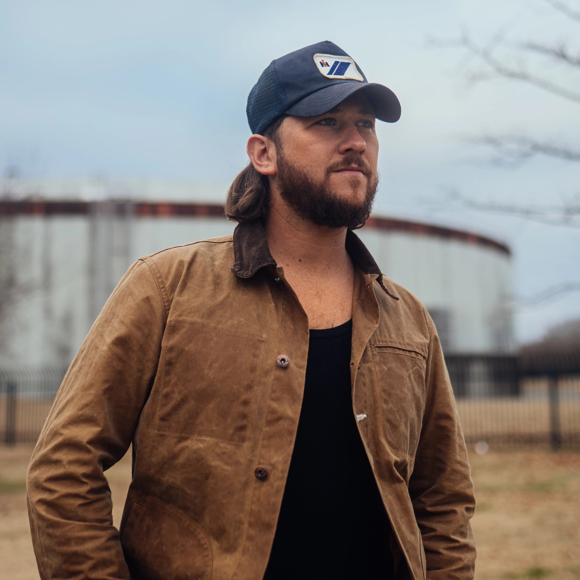 Bearded person in a brown jacket and cap stands near an industrial tank.