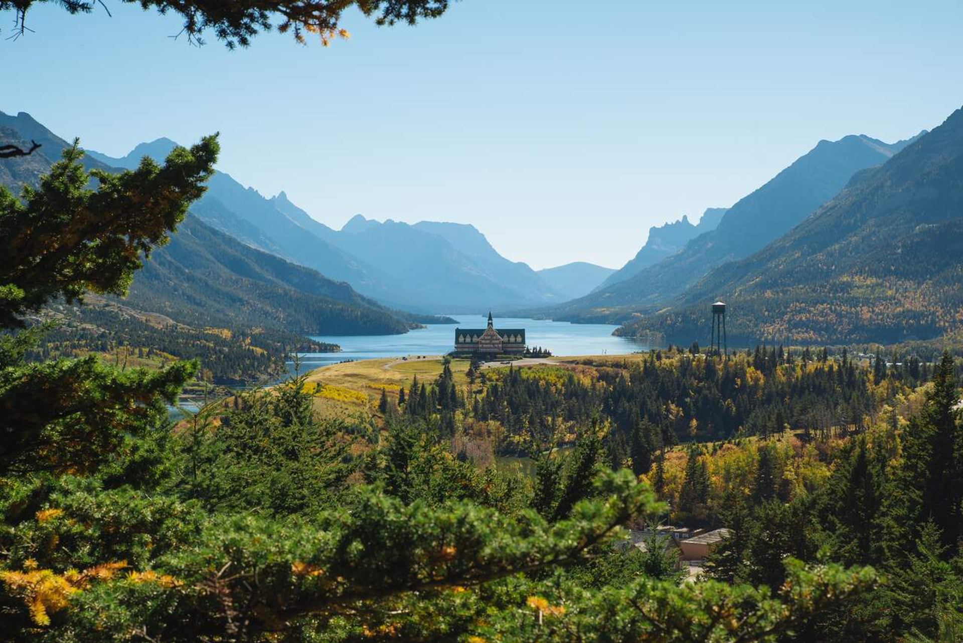 Mountain lake valley with forested slopes, distant lodge, and water tower under clear sky.
