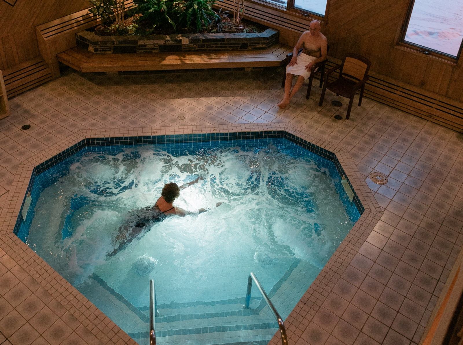 Indoor hot tub area with one person soaking and another seated nearby in a cozy lodge setting.