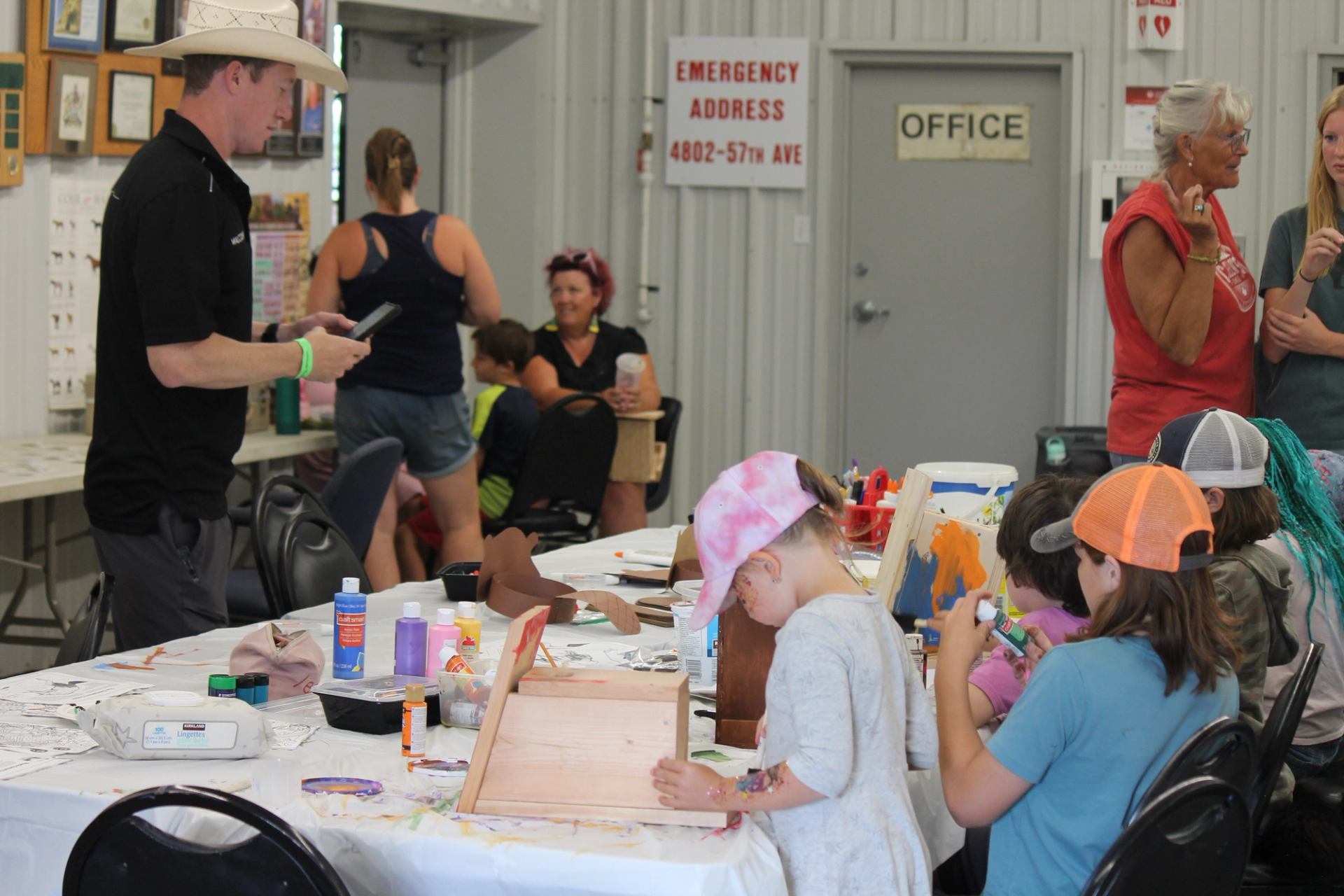 Children sit at a craft table painting wooden projects while adults supervise indoors.