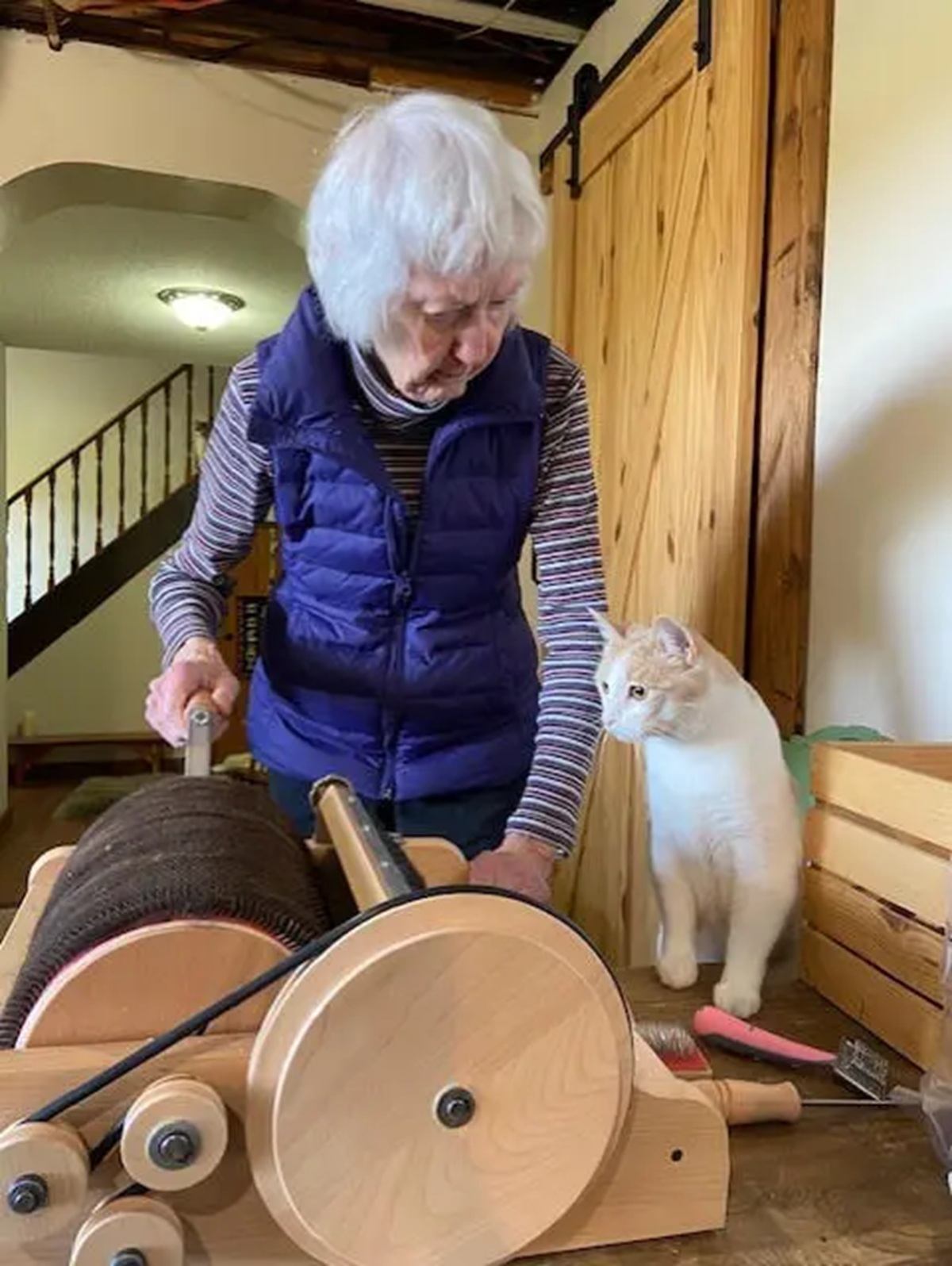 Person operating a wooden drum carder while a white cat watches on the table.