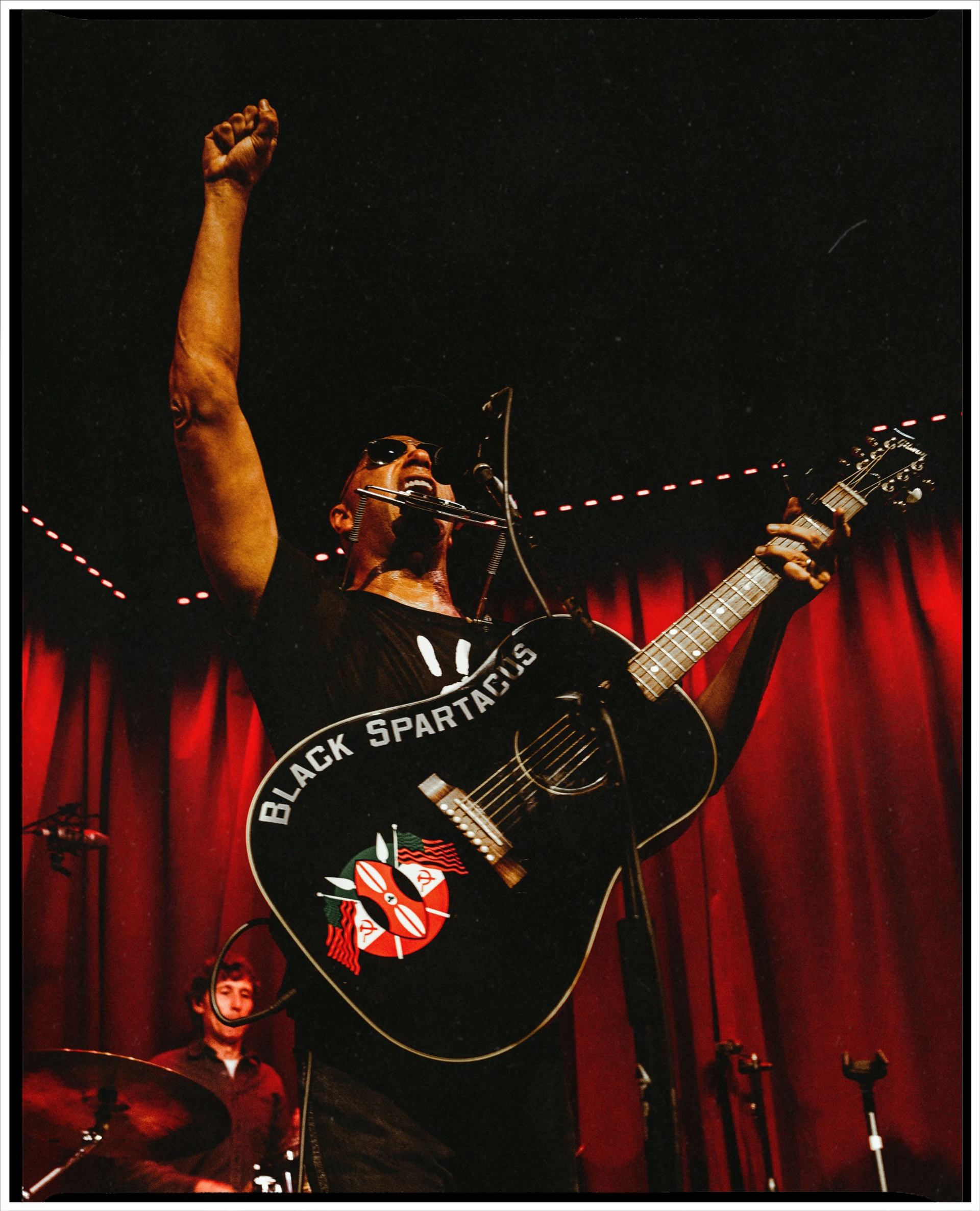 Musician onstage raises fist while singing and playing Black Spartacus guitar before red curtains.