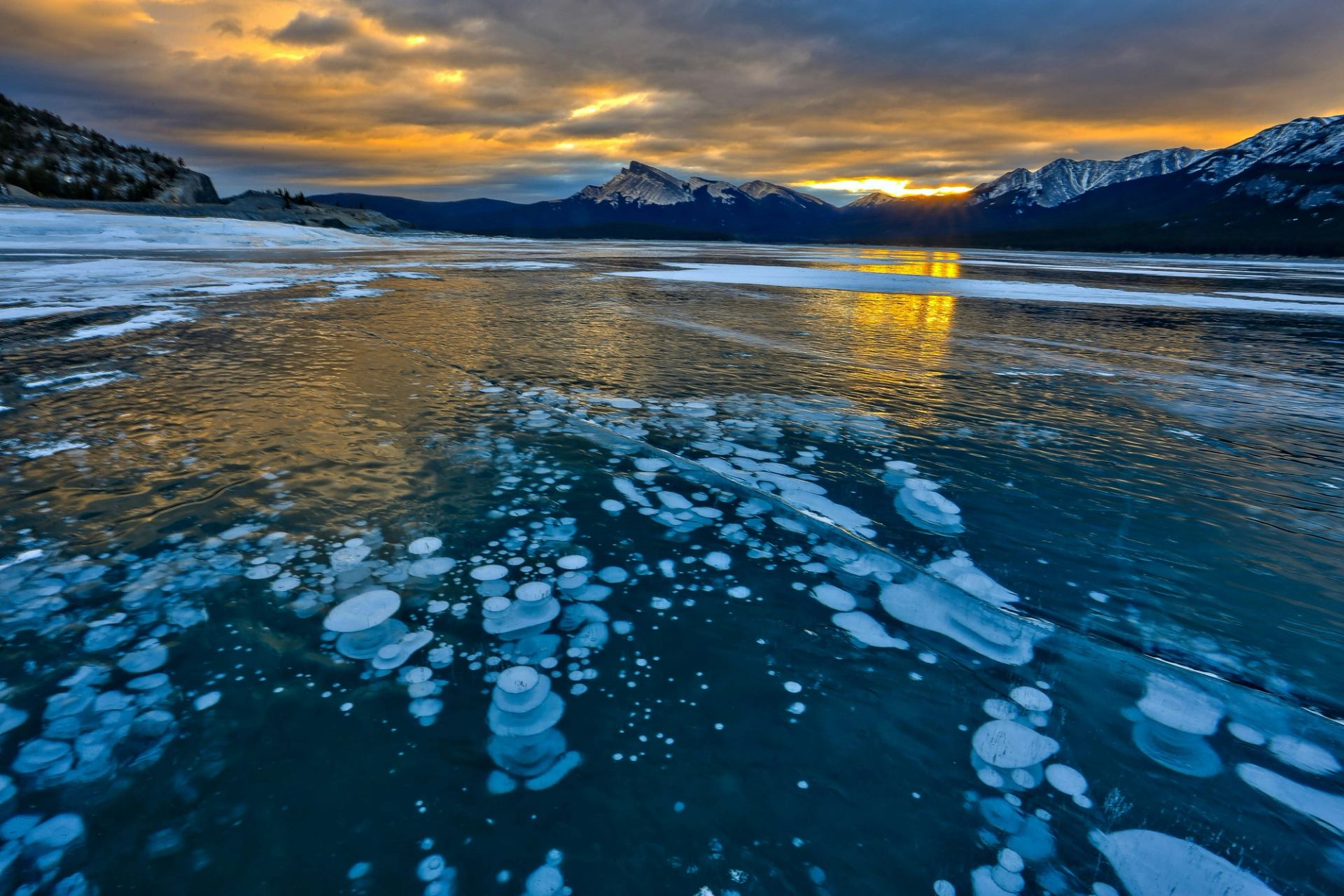 Frozen lake with trapped bubbles reflects a golden sunset beneath snow-covered mountains.