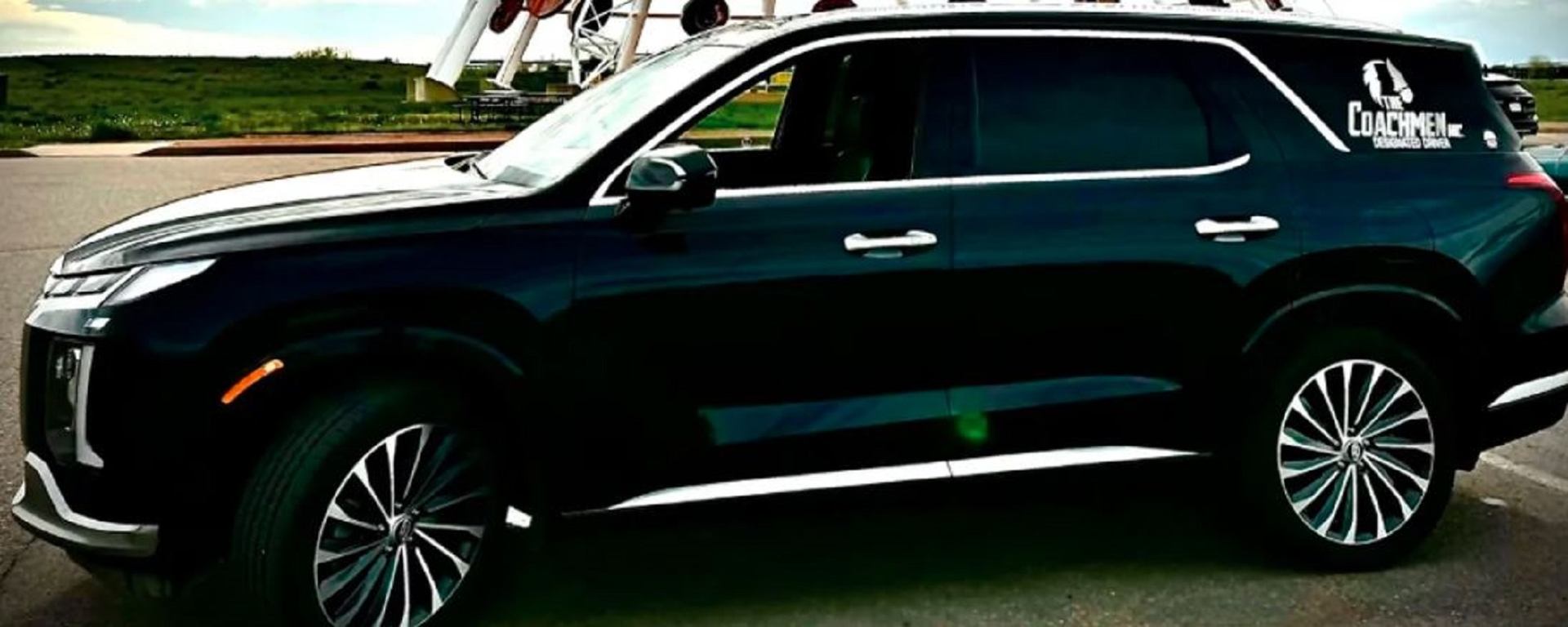 Black SUV with The Coachmen Inc. logo parked on pavement with greenery in background.