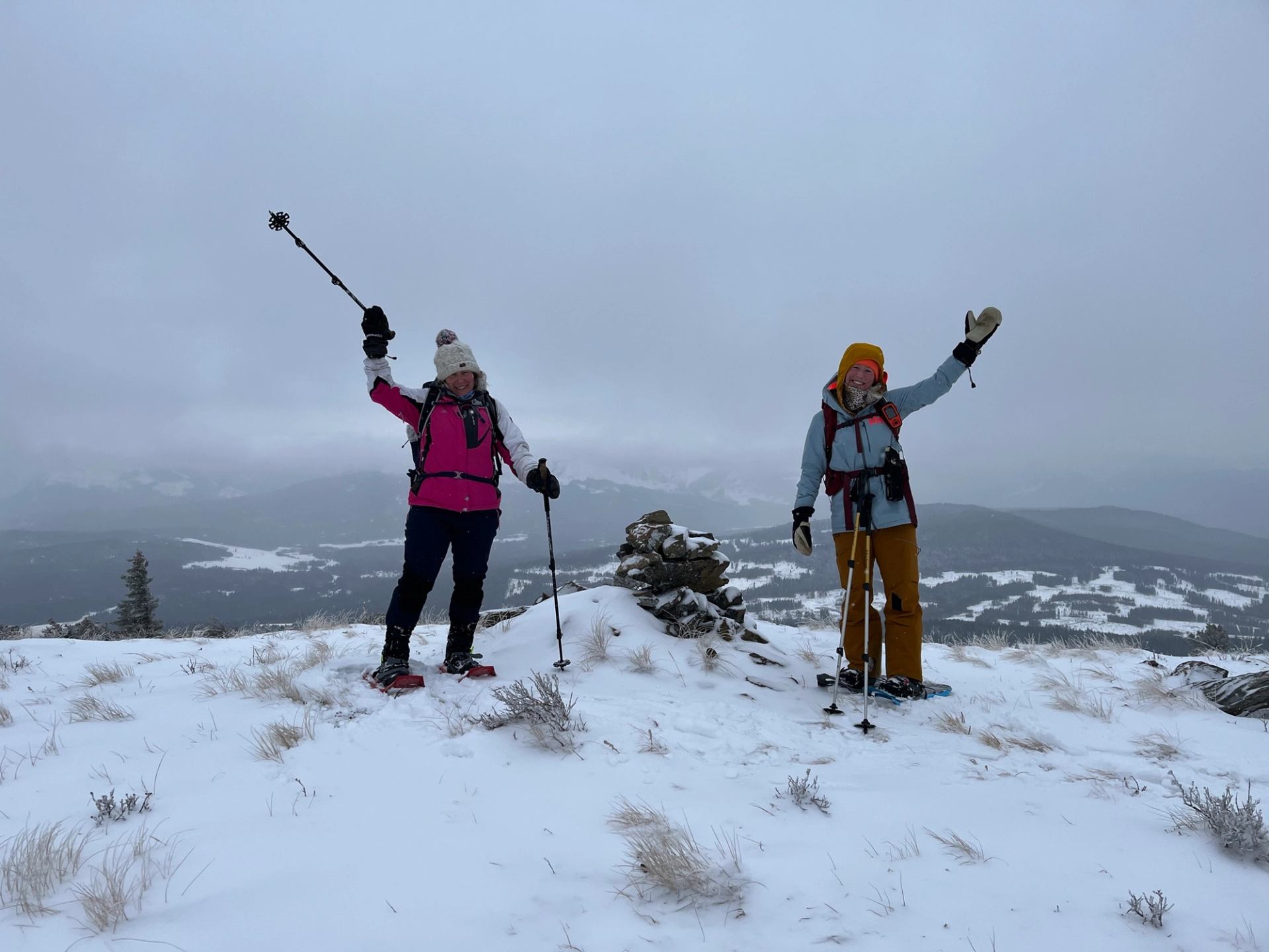 Two snowshoers standing on a snowy ridge, raising poles in celebration with cloudy mountains behind.